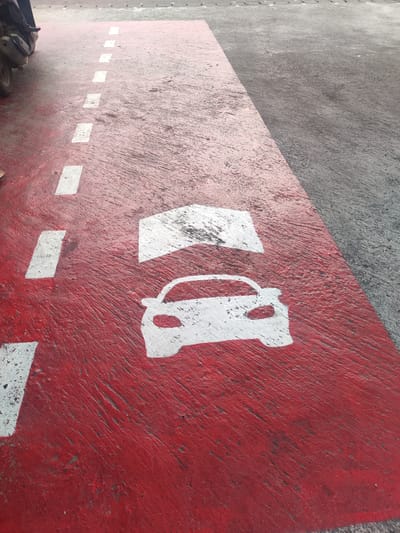 The image shows a designated car lane on a concrete surface in Bedulu, Indonesia. The lane is painted red and has white markings indicating the direction of car traffic, including a car symbol and an arrow. A series of dashed white lines form a lane marker. The lane is adjacent to a plain concrete surface. Part of a scooter is visible on the left. The scene is taken during the day. The concrete shows signs of wear and texture.