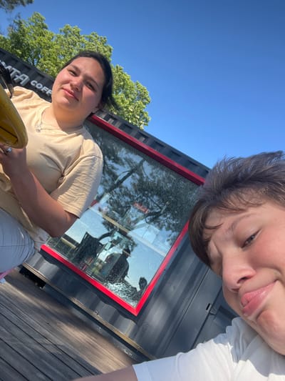 This media file features two young individuals, likely friends, posing for a selfie outdoors. The first person, in the foreground and to the left, is a young woman with dark hair pulled back and a light tan t-shirt. She is looking directly at the camera with a slight smile. She is wearing a necklace with a charm and a ring on her left hand. The second person, partially visible in the upper right of the frame, has brown hair and is looking down at the camera. They are wearing a white t-shirt.

The scene appears to be a sunny day, with a clear blue sky visible. They are standing near a dark, modular structure with a red trim around a large window. Through the window, reflections of trees and sky are visible, along with what appears to be a Coca-Cola logo and some interior elements, suggesting it might be a kiosk or a small building. The ground beneath them is made of wooden planks. The overall mood seems relaxed and happy, suggesting a casual outing.

Visible text includes "COFF" on the side of the dark structure, implying it may be a coffee stand or shop. The image appears to be taken in Ochamchire, Abkhazia. The sun is casting long shadows, indicating it might be late afternoon.