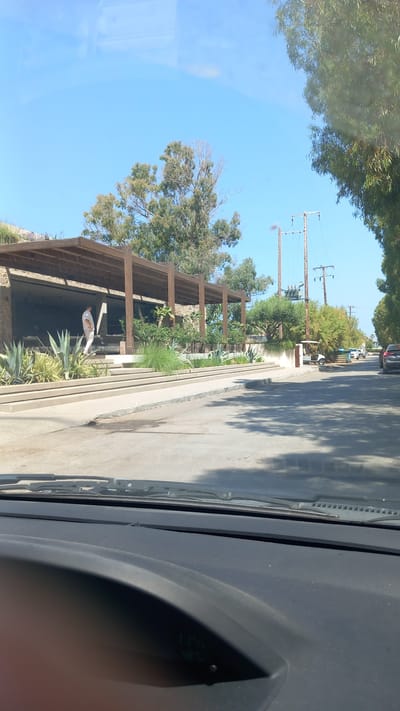 A person stands on a raised concrete walkway in front of a building with a wooden overhang in Lambi, Greece. The building appears to be a restaurant or similar establishment, with tables visible inside. Several large, spiky plants are arranged in front of the building. A paved road runs alongside the walkway, with a few parked cars and a golf cart visible further down the street. Trees and power lines are present in the background. The sky is clear and blue. The image is taken from the perspective of someone inside a vehicle, looking out through the windshield.