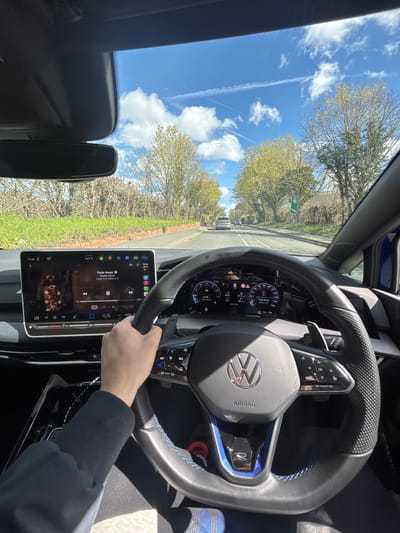 A photo, taken from the driver’s seat of a car, shows a person driving on a tree-lined road under a blue sky with contrails. The vehicle’s dashboard features a steering wheel with a Volkswagen logo, a digital speedometer, and a large infotainment screen displaying music information. The screen shows “Fade Away” by Roddy Ricch is playing. The speedometer reads 8 mph, and the dashboard shows 14.5 °C (58.1 °F). The photo may have been taken in or near Broxbourne, United Kingdom, given that a green road sign is partially visible in the background, which are commonly found in the UK. The location cannot be verified definitively with the available information.