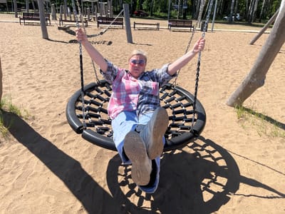 The image shows a woman sitting on a round swing in a sandy playground. She is wearing a plaid shirt, blue jeans, and white sneakers. Her arms are outstretched, and she is looking at the camera. The swing is made of black rope and has a black frame. In the background, there are trees, benches, and other playground equipment. The ground is covered in sand, with footprints visible. The lighting suggests it is a sunny day. The location is identified as a playground in Galewo, Russia.