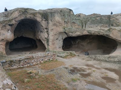 The image depicts the ancient cave city of Uplistsikhe, Georgia. The scene shows weathered rock formations with large cave entrances carved into the stone. In the foreground, there is a low stone wall constructed from various sized rocks, bordering an area with sparse, dry grass and patches of damp earth with puddles reflecting the overcast sky. The terrain is uneven and rocky, with some areas appearing smoothed by time and weather. On the top of the rock formations, several figures are visible from a distance, appearing as small silhouettes against the sky, engaged in exploration or observation of the site. One figure on the far left stands near the edge of a cliff. Another figure is positioned on a higher ledge to the right, seemingly engaged with a tripod, suggesting photography or surveying. A third, smaller figure is also visible on a distant ledge. The overall environment is natural and rugged, characteristic of an archaeological site.