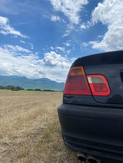 An extreme low-angle, eye-level shot captures the rear of a dark-colored sedan parked in a dry, grassy field with rolling, green mountains in the background. The sky above is a vibrant blue, dotted with fluffy white clouds. The car's taillight, a cluster of red and orange bulbs, is prominently displayed on the right side of the frame. The exhaust pipes are visible at the bottom of the bumper. The location is identified as Kurtovo Konare, Bulgaria.