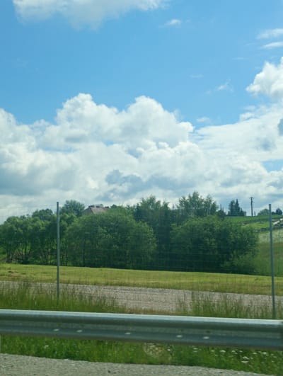 The image shows a rural landscape on a sunny day with a bright blue sky dotted with fluffy white clouds. In the foreground, a metal guardrail runs horizontally across the frame, separating a paved area from a grassy verge. Beyond the guardrail, a wide expanse of green grass stretches into the middle ground, interspersed with patches of gravel and tall, dry grasses. A wire fence runs through the middle ground, marking a boundary within the landscape. In the background, a dense line of trees forms a green barrier. Peeking above the trees is the roof of a building with reddish-brown tiles. Further in the distance, a utility pole with electrical wires is visible, indicating the presence of infrastructure. The scene suggests a quiet, outdoor environment, likely in a rural or semi-rural setting.