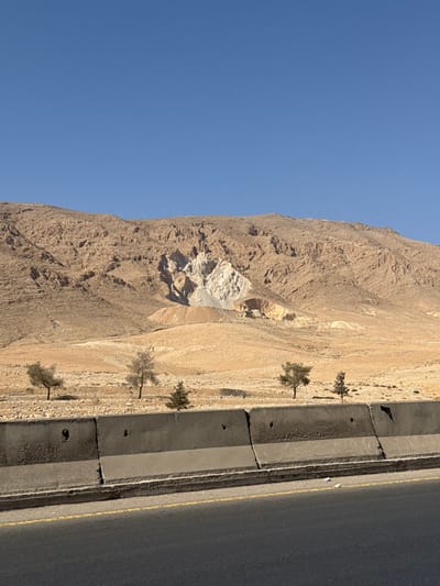The image depicts a desert mountain landscape under a clear blue sky, likely in the City of Adra, Syria. In the foreground, a dark asphalt road is visible, marked by a double yellow line. Immediately adjacent to the road is a weathered, segmented gray concrete barrier extending horizontally across the lower third of the frame. The barrier exhibits circular holes and some damage, including chipped sections. Beyond the barrier, a dry, rocky, and sparsely vegetated terrain slopes upwards towards a range of tan and light-brown mountains. Four small trees are present in this intermediate zone; two appear to be dormant deciduous trees, and two are small evergreens. On the central mountain slope, a significant white excavation site or quarry is visible, exposing lighter rock and featuring a grey spoil pile at its base. Faint, small, reddish-pink specks are discernible against the clear blue sky in the upper right portion of the image. No people or animals are visible.