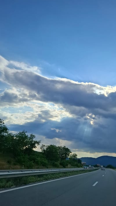 The image shows a view from a highway with vehicles on it. The highway has multiple lanes and a metal guardrail separating it from the side of the road. On the left side of the highway, there are green trees and vegetation covering a hillside. The sky is blue with dramatic clouds, and sunbeams are visible breaking through the clouds. In the distance, there are mountains. The scene appears to be a scenic view along a road in the city of Gorno Varshilo, Bulgaria.