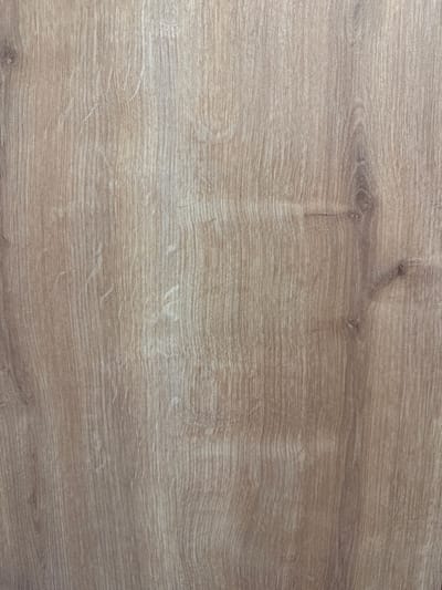 This is a close-up, full-frame photograph of a wood-grain texture. The wood is light brown with visible grain patterns running vertically. There are slight variations in color and tone throughout, with some lighter streaks and darker knots or imperfections. The texture appears smooth. There are no people, objects, actions, or interactions visible in the image. The location in the city of Gülnar, Turkey, is not ascertainable from the image itself.