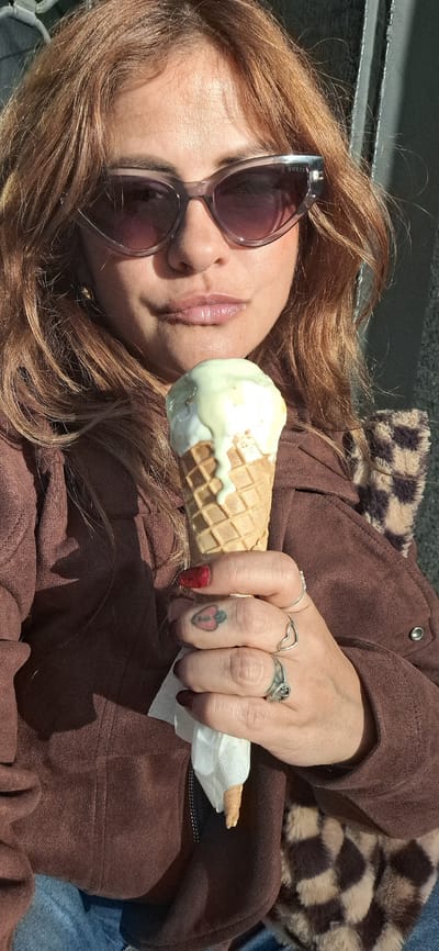 A woman with auburn hair and sunglasses is holding a melting ice cream cone in her right hand. The ice cream is light green and white, in a waffle cone. She has multiple rings on her fingers and a tattoo of a spade on her middle finger. She is wearing a brown jacket and blue jeans. A checkered, furry scarf or blanket is partially visible. The location is Santa Cruz De Tenerife, Spain, but specific location is not discernible from the image.