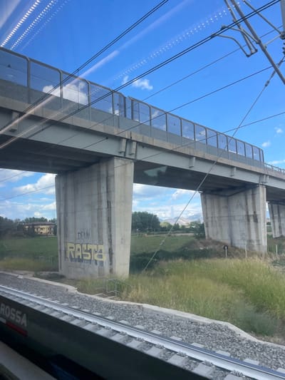 The image depicts a rural or semi-rural landscape observed from the window of a train, showing a large concrete overpass structure consistent with a high-speed railway line near Afragola, Italy. The sky is bright blue with scattered white cumulus clouds. Multiple overhead electrical catenary wires span across the upper portion of the frame, showing reflections of interior train lights as parallel rows of white dots.

In the midground, a substantial concrete overpass is the dominant feature, supported by multiple robust concrete piers. The overpass deck features a gray metal safety barrier or fencing along its edge. The concrete piers exhibit weathering and dark vertical runoff stains. On the face of the leftmost visible pier, yellow graffiti spells "RASTA" with smaller black text "DEAN" positioned above it.

The ground beneath the overpass is covered in green and dry brown grasses and low vegetation. To the left, in the mid-distance, a multi-story building with a terracotta-tiled roof and light-colored walls is partially visible amidst trees. To the right, beyond the overpass piers, distant mountains or large hills are discernible under a light haze. A small set of concrete steps with metal handrails is situated at the base of the graffiti-marked pier.

In the foreground, the image captures a portion of parallel railway tracks composed of concrete sleepers and gravel ballast. The lower left corner of the frame reveals the dark gray exterior of the train from which the photograph was taken, with the partial word "ROSSA" visible in white lettering, indicative of a high-speed train operating in Italy. No individuals are visible in the scene. The overall impression is one of infrastructure traversing natural and built environments.