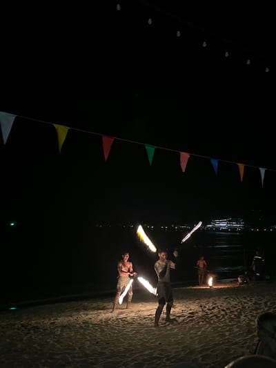 The image depicts a nighttime scene on a sandy beach, likely in Hat Khai Muk, Thailand. Three individuals are performing fire dance with flaming batons near the water's edge. The beach sand appears textured and lit from the fire.  In the background, there are decorations including pennant flags, lights and the water.  The scene is overall dark, with illumination primarily from the fire.