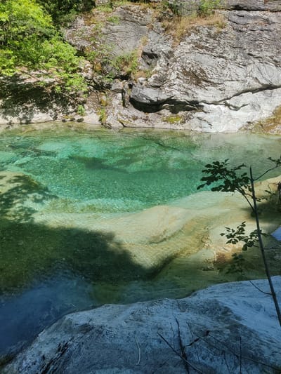 A clear, turquoise river flows through a rocky gorge. Sunlight dapples the water's surface, illuminating the smooth, submerged stones below. Lush green foliage spills over the rocky banks, casting shadows on the water and the surrounding landscape. The scene is tranquil and natural, suggesting a secluded spot within the Municipal Unit of Melivia, Greece.