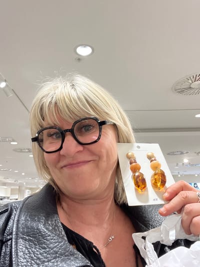 A woman with blonde hair and glasses displays a card with two amber-colored beaded hair clips. She wears a black leather jacket and a black top with a delicate silver necklace. The background appears to be a store, indicated by ceiling lights and displays. A white shopping bag is visible in her hand.