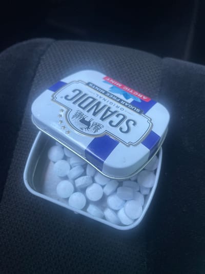 The image shows an open tin of Scandic Arctic Mint sugar-free mints. The tin is predominantly white with blue stripes and features a logo of a moose or elk. The text "SCANDIC", "ORIGINAL", "SUGAR FREE MINTS", and "ARCTIC MINT" is visible on the tin. The tin is filled with small, round, white mints. The tin is placed on a dark, textured surface, possibly a car seat. The lighting suggests it could be daytime, with natural light illuminating the mints. There are no people or animals visible in the image. The scene appears to be casual and everyday. The provided location context is Ganderkesee, Germany.