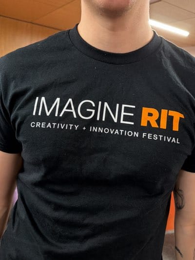 A person is wearing a black t-shirt with the words "IMAGINE RIT" in white and orange font, and below it, "CREATIVITY + INNOVATION FESTIVAL" in white font. The setting is likely indoors, possibly at the Rochester Institute of Technology (RIT) in Rochester, United States, based on the t-shirt's logo. The person is standing with a portion of their upper body visible, including part of their neck and shoulder. There is some other coloration visible.