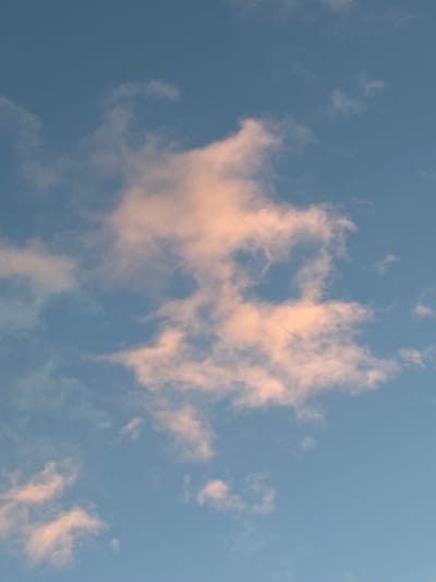 A high-angle, portrait-oriented, full-shot image captures a pale blue sky with scattered clouds tinged with pink from what appears to be a sunset or sunrise. The clouds are soft and wispy, with some denser formations appearing in the upper center and right of the frame, while thinner wisps are visible towards the bottom. The lighting suggests a warm, diffused glow, creating a serene atmosphere. No people, structures, or earthly elements are visible; the image is solely focused on the sky. This scene is from Voeikovo, Russia.