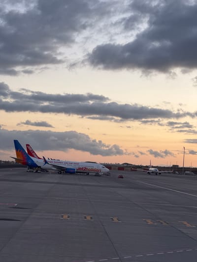 The image shows a Jet2 Holidays airplane on the tarmac at Tenerife South Airport (TFS) in Granadilla de Abona, Spain. The plane is white with blue and orange accents, including the Jet2 logo on the tail and fuselage. The sky is filled with clouds illuminated by a setting or rising sun, creating a warm glow. The tarmac is grey and marked with yellow lines and painted markings. Other airplanes are in the background along with some vehicles and airport infrastructure. There is no sign of people.