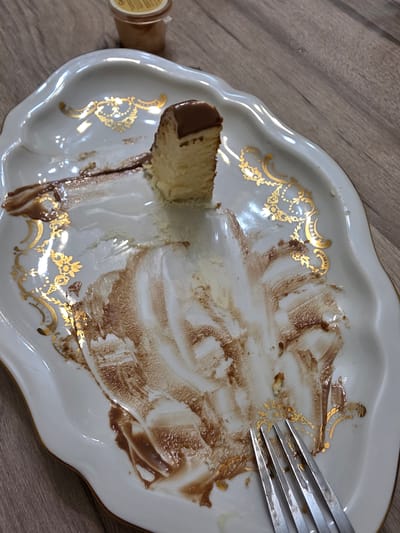 An overhead, slightly angled shot shows a slice of cheesecake with chocolate frosting on a white, ornate plate. The plate is dirty with chocolate smears and crumbs. A metal fork with four tines rests on the plate in the lower right corner. In the upper portion of the image, a small plastic container with a yellow lid is visible. The setting is a wooden surface. No specific location in Yalangach, Uzbekistan, can be determined from the image.