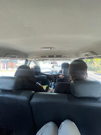The image shows the interior of a vehicle, likely an SUV, traveling along a road in Kalabbirang, Indonesia. Four individuals are visible. In the foreground, two people are seated in the back row, facing forward. The person on the left wears a dark hooded garment, and the person on the right has dark hair tied back. In the middle row, two individuals are seated. The person on the left, in the driver's seat, appears to be wearing a patterned shirt. The person on the right is looking towards the front and holding a mobile phone. The vehicle's interior includes black leather seats, air conditioning vents on the ceiling, and handles. Outside the vehicle's windows, the scenery suggests an urban or semi-urban environment with buildings and vegetation. The lighting indicates it is daytime.