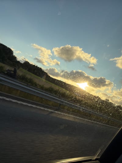 The image is a tilted, angled shot taken from inside a vehicle, likely on a highway, with the sun setting or rising. The foreground shows a dark asphalt road with white lane markings, blurred by motion. A metal guardrail runs along the side of the road, separating it from a grassy embankment covered with vegetation. Beyond the embankment, a dark, wooded hillside rises, with trees silhouetted against a bright sky. The sky itself is a mix of clear blue and scattered, fluffy clouds illuminated by the warm, golden light of the sun, which is positioned behind some of the clouds, creating a halo effect. The light also reflects off the car's windshield, creating a speckled pattern. In the distance, on the left side of the road, a dark, silhouetted figure on a motorcycle is visible, also blurred by motion. The overall scene suggests a journey on a highway near Ödsmål, Sweden, during a scenic time of day.