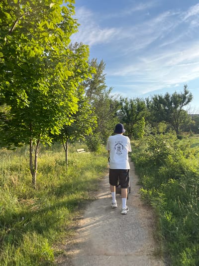In a sun-drenched, grassy park in Kotovo, Russia, a person walks away from the camera on a dirt path. They are wearing a white t-shirt with graphics on the back, black shorts, white socks, and athletic shoes. A dark blue bucket hat covers their head. The person is pushing a stroller. The park is filled with lush green trees and foliage, with a bright blue sky dotted with wispy white clouds overhead. In the background, other trees and greenery are visible, with faint hints of buildings in the distance. The sunlight casts long shadows across the path, indicating it is late afternoon or early evening.
