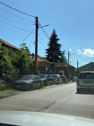 In Beli Osam, Bulgaria, a street scene unfolds under a clear blue sky. Several parked cars line the paved road, which has visible cracks and wear. The vehicles include a dark gray BMW coupe with license plate "OB 7727 BT" parked on the left, followed by a silver hatchback, another silver car, and a light green minivan with license plate "OB 9023 BH" further down the road.
On the left side of the street, houses with red-tiled roofs are visible, partially obscured by lush green trees and bushes. A large, dark green evergreen tree stands tall in the background. Utility poles with numerous wires crisscross the sky, and streetlights are mounted on some of them. In the distance, rolling green hills form the backdrop. Two flags are visible hanging from one of the houses, one appears to be the Bulgarian flag. The scene appears to be a quiet residential area.