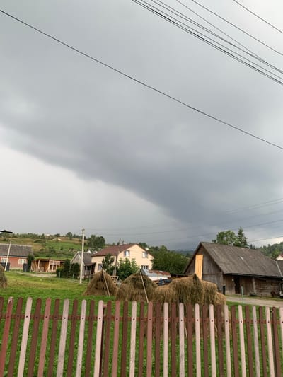 The image displays a rural landscape in Lazeshchyna, Ukraine, under a heavily overcast, dark grey sky. In the foreground, a wooden picket fence, featuring vertical slats painted in alternating light brown, dark brown, and faded off-white/green, extends across the frame. Immediately behind the fence, a green grassy field contains several conical haystacks.

The mid-ground is occupied by multiple structures. On the right, a large, dark wooden barn with a corrugated roof is prominent; its main entrance is dark, and a lighter-colored wooden board is affixed to its left side. To the left of the barn, residential buildings include a two-story light beige house with a brown roof, a smaller older dwelling, and a brick house (possibly red) exhibiting exposed wooden framing, suggesting ongoing construction or renovation. Multiple utility poles and numerous overhead lines traverse the scene horizontally.

The background consists of undulating green hills, indicative of a mountainous or hilly terrain. The sky is dominated by a massive, low-hanging, dark grey cloud formation, consistent with a storm front or heavy rain clouds. No individuals are present in the depicted scene.