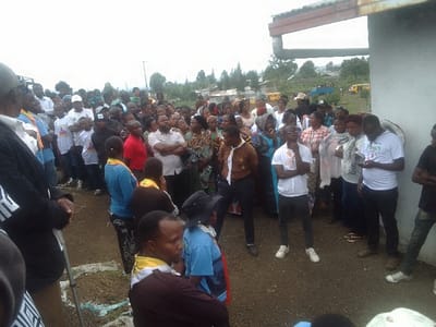 A large crowd of people is gathered outdoors near a building. Many wear white shirts, some with visible logos. Several individuals are adorned with yellow scarves or neckerchiefs, suggesting a possible scouting or youth organization connection. In the background, there is a landscape with houses and vegetation. School buses are visible. The setting appears to be a community event or gathering.