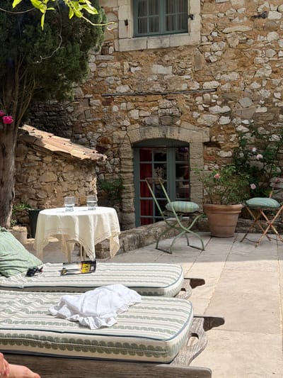The image depicts an outdoor setting in Saint-Victor-la-Coste, France. The scene includes stone walls, a window with red curtains, and chairs with cushions. A table with a white lace tablecloth stands nearby. Also visible are two books, a white shirt, a pair of keys, and a foot.