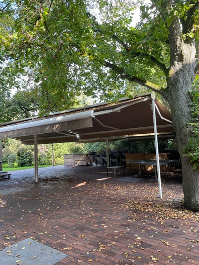 The image displays an unoccupied outdoor paved patio area in La Wantzenau, France, situated under a large, extended brown canvas awning supported by white metal poles. The top surface of the awning and its white gutters are partially covered with scattered fallen leaves. The ground is paved with reddish-brown rectangular bricks, extensively covered with fallen yellow and brown deciduous leaves, particularly toward the right and center. A section of gray concrete is visible in the immediate foreground.
On the right side of the frame, a large tree with a textured trunk and green foliage stands next to the awning, its branches extending over the structure. Underneath the awning, to the right, several stacks of dark-colored, metal-framed chairs are visible, alongside multiple round, metal-topped tables with central support pedestals. Towards the rear-left, a stack of light-colored wooden crates or pallets is present, adjacent to a simple wooden bench. The scene is illuminated by natural daylight, casting shadows from the trees and awning supports. No individuals are present, and no activities or interactions are depicted. The environment appears to be in an inactive state, potentially a seasonally closed or currently unused outdoor dining or gathering space.