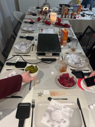 The image captures an overhead view of a long dining table set for a festive raclette dinner, likely in an indoor home setting in Bellevue, Switzerland.
**Subjects:** Two sets of hands are visible: one placing a white bowl filled with small green pickles (cornichons) onto the table, and another holding a mobile phone above an individual raclette pan. No full people are shown.
**Objects:** The table is covered with light-colored placemats and is extensively decorated. A large, black and silver raclette grill is centrally positioned. Numerous white rectangular and square plates are set, each accompanied by black cutlery (knives, forks, raclette spatulas). Paper napkins with a design featuring a white sphere and silver baubles are placed on some plates. Bowls of food include a large white bowl heaped with thinly sliced red cured meat and the aforementioned bowl of pickles. Various glasses, some containing an amber beverage, are scattered across the table.
**Christmas Decorations:** The setting is adorned with Christmas decorations, including a small, artificial Christmas tree with red baubles and lights, white decorative balls resembling snow, and artificial snowflakes sprinkled on the table. Two white candles in glass holders provide ambient light. Santa Claus-themed cutouts or placemats add to the festive atmosphere. Small gift boxes and spice racks are visible at one end of the table.
**Activity/Event:** The arrangement clearly indicates a raclette dinner, a communal and interactive meal, suggesting a social gathering for friends or family. The Christmas decorations imply this is a holiday celebration.
**Notable Details:** The indoor lighting suggests the event is taking place in the evening or at night. The overall impression is one of a warm, festive, and convivial occasion.
**Visible Text:** The word "CHRISTMAS" is prominently displayed on a white card near the raclette grill. A small white name card with "Calou" written on it is visible near a wooden tree-shaped ornament.
