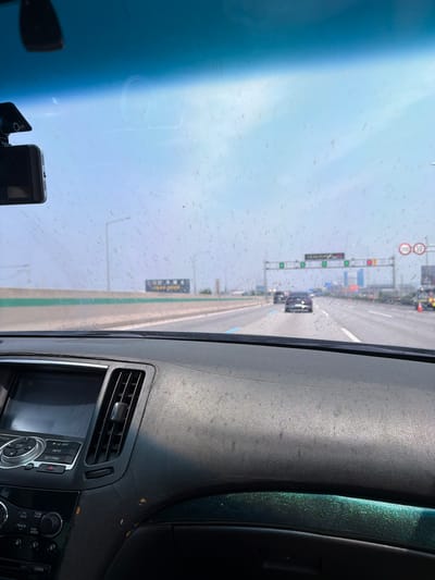 The image is taken from inside a car, looking out through the windshield, which is covered in numerous small, brown specks, likely dust or dirt. The car appears to be driving on a highway. In the foreground, the dashboard of the car is visible, with a navigation screen, air vents, and various buttons. Beyond the windshield, the highway stretches into the distance, with other cars visible. There are overhead signs and traffic lights, indicating an urban or suburban environment. The sky is overcast or hazy. Based on the context of the highway infrastructure and the visible signage in the distance (which resembles South Korean road signs, though not fully legible), this scene is likely located in or near Anseong-si, South Korea.