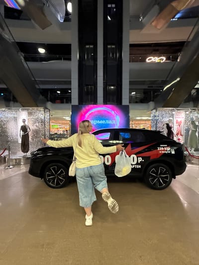 A woman stands in front of a black car in a shopping mall. She is wearing a yellow long-sleeved shirt and light blue wide-leg jeans. She holds a white plastic bag containing yellow objects in her right hand and extends her left arm towards the car. The car has red and black graphics on its side, with text that reads "Звони 239-939 и получи 100000р. АРТЕН". In the background, there are displays with mannequins, a large digital screen displaying neon pink text, and escalators. The scene appears to be set in a shopping mall in Ermakovo, Russia, based on the text on the car, which is a Russian phrase referring to a promotion.