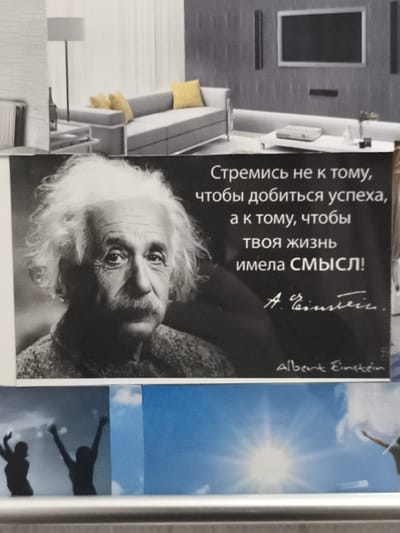 The image shows a collage of various photographs. The top section depicts a modern living room interior with a sectional sofa, yellow cushions, a floor lamp, and a large television mounted on a wall. The central section features a close-up portrait of Albert Einstein with a quote in Russian superimposed. The quote reads, "Strive not for success, but for a life that has meaning!" followed by Einstein's signature. The bottom section displays silhouettes of two figures with their arms raised against a sky background, and another image of a bright sun in a blue sky. There is no information or visual evidence to suggest the location is Eitorf, Germany.