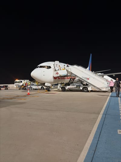 An airplane with the logo "AP AVIAPARTNER GO GREEN" is parked on the tarmac at night. A mobile stairway is attached to the aircraft, and two people are descending it. Baggage carts and service vehicles are visible in the background, and an orange traffic cone is placed on the tarmac. The scene appears to be at an airport.