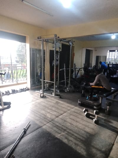 The image displays an indoor gym facility situated in Njoro ward, Kenya. The interior features light-colored walls and ceiling, with an activated linear LED light fixture mounted on the ceiling in the foreground. The floor is covered with dark grey interlocking rubber mats, across which distinct parallel shadows are cast from a large, open entryway on the left side of the frame. This opening, equipped with a metal railing, offers a view of an exterior with visible vegetation and overhead power lines. A row of approximately ten pairs of assorted athletic shoes is neatly aligned against the wall next to the opening.
In the center-left, a silver multi-functional cable machine with pulleys and various attachments stands prominently. A silver barbell lies diagonally on the floor in the immediate foreground. Towards the mid-right, a male individual, appearing light-skinned and wearing a light-colored long-sleeved shirt and dark shorts with yellow accents, is seated on a black adjustable weight bench. His back is partially oriented towards the viewer, and his hands are positioned in his lap. Adjacent to the bench on the floor are a black aerobic step platform and two hexagonal dumbbells.
Further into the gym, in the background, a second male individual dressed in a blue long-sleeved shirt and dark pants is observed standing near additional weightlifting equipment, appearing to be bending over. The rear wall features a large black banner or poster with white text, partially obscured, and several mirrors reflecting sections of the gym. A window with a grid pattern is visible on the far right background. The area deeper within the facility is noticeably darker than the brightly lit foreground, indicating reliance on internal illumination and ambient light.