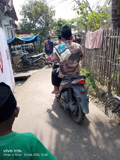 A man in a patterned shirt and black cap is riding a scooter down a narrow street. He has a prayer rug draped over his shoulder. Other men in similar attire walk ahead of him. To the left, a motorcycle is parked next to a food cart. A fence runs along the right side of the street, with a towel hanging on it. A boy in a green shirt and a black cap is partially visible in the foreground.