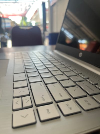This media file is a close-up, angled shot of a silver laptop keyboard. The laptop appears to be open, with the screen on the left side of the frame, reflecting a blurry, out-of-focus scene that includes bright lights and possibly some people. The keyboard itself is the primary focus, with many keys clearly visible, including "Insert", "backspace", "pg up", "enter", "pg dn", "pause", "shift", "end", "ctrl", and arrow keys. The keys are clean and appear to be in good condition.

The lighting suggests it might be daytime, with natural light contributing to the reflections on the screen. The background is very blurred, making it difficult to ascertain the exact setting, but the reflections on the screen hint at an indoor environment with some activity or decoration. There are no people directly visible in the frame. The overall impression is that of a snapshot of a workspace, possibly taken during a break or while working. The visible text includes labels on the keyboard keys and the brand logo on the laptop's bezel.