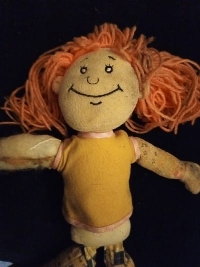 A well-loved, handmade cloth doll is centered. It has a round, beige head with simple, stitched facial features: arched eyebrows, round eyes, a curved nose, and a smiling mouth. The doll's hair is vibrant orange yarn, styled in a messy, playful way. It wears a yellow shirt with pink trim. Its arms are made of patterned fabric, and one has a tear revealing the stuffing. The doll has checkered plaid pants. The background is dark, providing a contrast to the doll's colors.