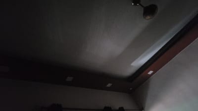 The image shows a ceiling in a room in Kanpur, India. There is a ceiling fan and a brown border along the top of the wall. There are two light fixtures on the ceiling. The room is dark and there are no people in the image. The coordinates provided are for Kanpur, India. The image appears to be taken from the corner of the room, looking up at the ceiling. The ceiling is painted in a light color, and the brown border is a contrasting color. The fan is in the center of the image, and the light fixtures are on the sides. The bottom of the image is out of focus, suggesting that the image was taken from a low angle. The room appears to be empty.