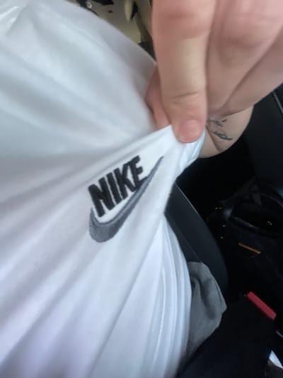 A close-up, slightly blurred shot shows a white t-shirt with a black Nike logo. The t-shirt is being held by a hand, with part of a tattoo visible on the forearm. In the background, a black bag and a red object are visible. The scene appears to be inside a vehicle. There is no information about the location in Kipra, Bulgaria.