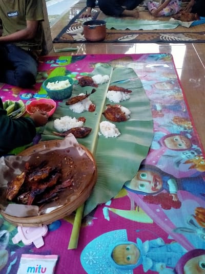 The image captures an informal communal meal in Celep, Indonesia, with participants seated on floor mats. A large banana leaf, serving as a primary eating surface, is centrally positioned on a vibrant pink mat adorned with cartoon characters. On the banana leaf, approximately six individual portions of white rice are visible, each accompanied by a piece of grilled fish and a small amount of red chili sauce (sambal). In the lower-left foreground, a circular woven bamboo basket, lined with brown paper, holds additional grilled fish. Adjacent to this, a person wearing a dark green long-sleeved shirt has a hand near a pink plastic bowl containing red chili sauce, next to which a small dish of sliced green vegetables and a light blue plastic basket with white food items are placed. In the upper-left, another individual in a dark green "Maternal" t-shirt and dark pants sits on a darker, patterned floor mat, near a lidded bronze pot. A child's leg is partially visible in the upper-right. The floor consists of tiled sections under the mats. A "mitu" packet and a small pink plastic object are visible on the pink mat in the lower-left.