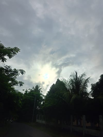 The image shows a cloudy sky above a road in Meunasahteungkudigadong, Indonesia. Trees are visible along the roadside, with a few palm trees interspersed. A fence runs alongside the road. The sun is obscured by the clouds, but its position is indicated by a bright patch in the sky. A person on a motorcycle can be seen on the left side of the road.