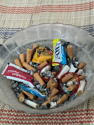 A clear, scalloped glass ashtray, heavily soiled with cigarette ash and numerous discarded cigarette butts, is centrally positioned on a woven mat. The mat displays a pattern of horizontal stripes in beige, white, red, and blue. Inside the ashtray, approximately 30-40 cigarette butts are visible, mixed with dark grey ash; some butts show white filters and partial brand names. Interspersed with the butts and ash are various items of refuse: a yellow and blue "FOX'S CRYSTAL CLEAR LEMON MINT" candy wrapper, a red and white "Sambal Pedas" sachet, a crumpled yellow and orange snack wrapper, a small blue foil wrapper, two red rubber bands, and a small, irregular white plastic object. The scene is located in Kumbang, Indonesia.
