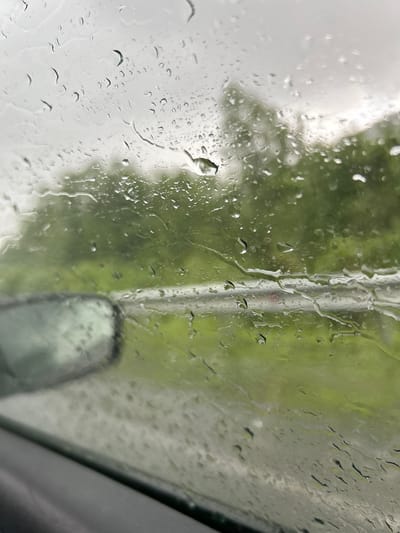 The image shows a view through a rain-streaked window, likely a car window, looking out at a blurred green and gray landscape. Water droplets of various sizes are visible on the glass, distorting the view of trees and a road. A dark, out-of-focus side mirror is visible on the left side of the frame. The sky appears overcast. The scene suggests a rainy day in an outdoor environment, possibly on a road trip or commute. The location in the city of Кулмас, Russia, is not discernible from the image itself.