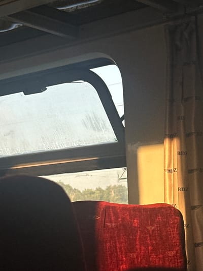 The image depicts the interior of a train carriage. The foreground features two red velvet seats, with the back of one seat prominently visible on the left, and the headrest of another on the right. The window, partially open, reveals a hazy view of trees and foliage outside, suggesting a landscape setting. Sunlight streams in from the right side of the carriage, casting a warm glow on the wall and a beige curtain patterned with the letters "BDZ" and a winged emblem. The ceiling of the carriage is visible at the top, with visible lighting fixtures. This scene is consistent with the interior of a Bulgarian State Railways (BDZ) train traveling through the Bulgarian countryside, possibly near Tulovo, Bulgaria, a town known for its railway infrastructure.