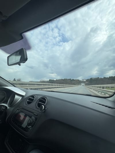 The scene is captured from the interior of a vehicle, facing forward through the windshield on an overcast day. The vehicle's black dashboard, including two circular air vents on the passenger side, a central infotainment unit with a black screen and control knob, and a magnetic phone holder mounted on an air vent, are visible in the foreground. A black-framed rearview mirror is positioned at the top center of the windshield.

Outside the vehicle, a two-lane roadway, appearing to be an elevated section or bridge, extends into the distance. Metal guardrails line both sides of the road. The surrounding landscape consists of mostly barren deciduous trees and some evergreen foliage on either side of the road, suggesting late autumn or early spring. Several distant white vehicles are visible on the road ahead, traveling in the same direction. A cluster of white structures or buildings can be faintly observed on a distant elevated area to the right of the road. The sky is predominantly covered by grey and white clouds. No individuals are visible. The specific location within the city of Kumanovo, North Macedonia, cannot be determined from the visual evidence presented in the image.