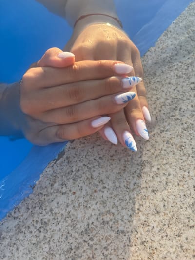 This is a close-up, overhead shot of a pair of hands emerging from blue water, resting on a speckled, granite-like surface. The hands have almond-shaped, white nail polish, with some nails featuring delicate blue floral designs. The water is bright blue and appears to be shallow, with sunlight reflecting off its surface. The granite surface is textured with small specks of various colors, including gray, brown, and black. The image captures a moment of relaxation, likely by a swimming pool or a body of water. The location in the city of Delchevo, North Macedonia is not discernible from the image itself.