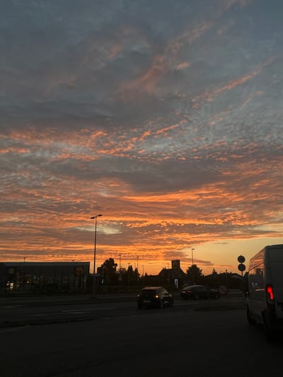 This image depicts a street scene at sunset in Snoghøj, Denmark. The sky is filled with dramatic clouds illuminated by the setting sun, displaying vibrant shades of orange and pink against a darker gray backdrop. In the foreground, a dark asphalt road is visible, with a few cars driving. To the left, the facade of a Lidl supermarket is apparent, with its illuminated signage. Several streetlights are also visible throughout the scene, some casting long shadows. The overall impression is of an urban environment during twilight, characterized by a striking natural sky display.