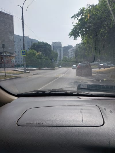 A car's interior perspective shows a street scene in Belokurikha, Russia, on a bright, hazy day. The view is through a windshield covered in dust and streaks. On the left, a tall, grey brick building dominates the background, with a smaller, lighter building with multiple windows next to it. A pedestrian crossing sign is visible on the left side of the road. Trees line the right side of the street, their leaves creating a dappled effect. Several cars are on the road: a dark SUV in the foreground, a lighter car behind it, and another dark sedan further down the road. The road itself appears to be asphalt, with lane markings visible. A bus stop sign is also present on the right side of the road. The dashboard of the car is visible in the foreground, with a textured surface and a compartment with a slight sheen.