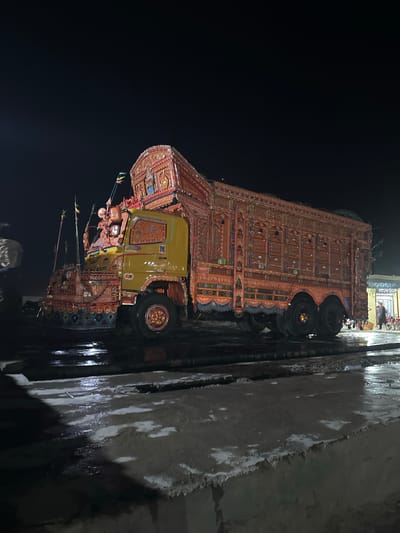 An elaborately decorated, bright yellow and orange truck is parked at night on a wet, paved surface. The truck is adorned with intricate patterns, colorful artwork, and multiple flags. The rear of the truck features a large, multi-wheeled cargo bed also covered in similar detailed decorations. In the background, a dimly lit building with some signage is visible, and a person can be seen standing near it. The overall scene is illuminated by artificial light, creating reflections on the wet ground.